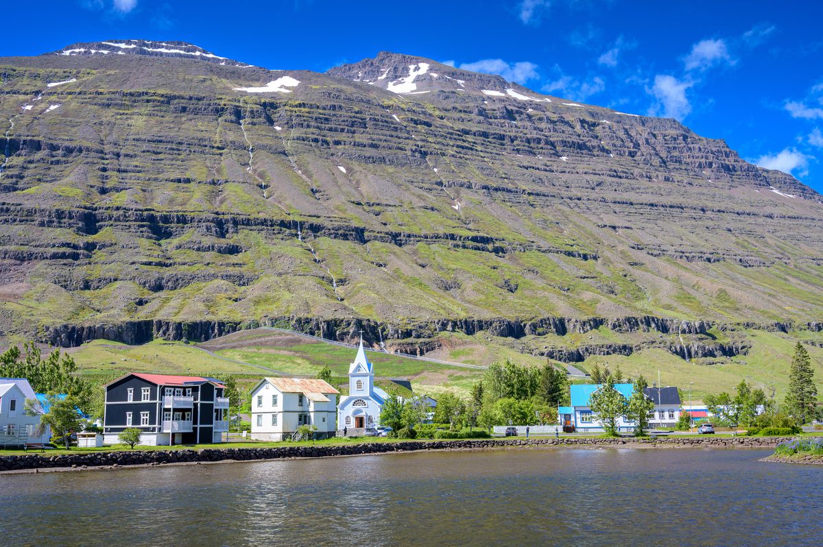 Colorful waterfront buildings and the pale blue church sit across still water beneath a towering banded mountain.