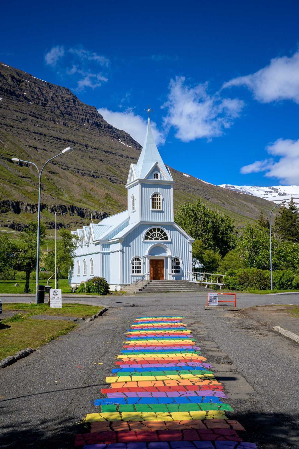 Seyðisfjörður's rainbow-painted street leads directly to the pale blue church under a brilliant sky.