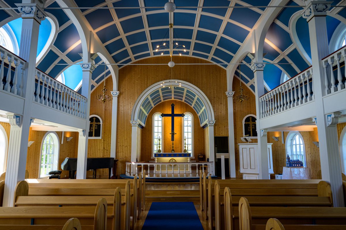 The blue-and-cream interior of Seyðisfjörður's church frames wooden pews, a central aisle, and a glowing cross.