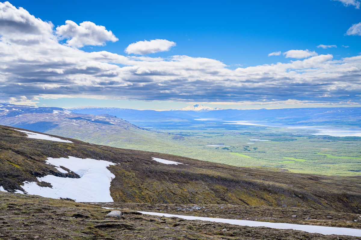 Patchy snow and a bare roadside foreground overlook the broad green plains and distant mountains of Hérað.