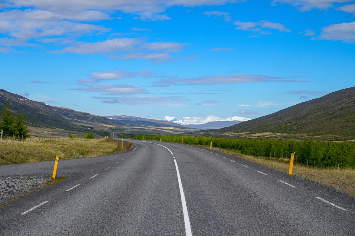 An empty paved road runs between green hills toward a distant valley beneath high blue summer clouds.