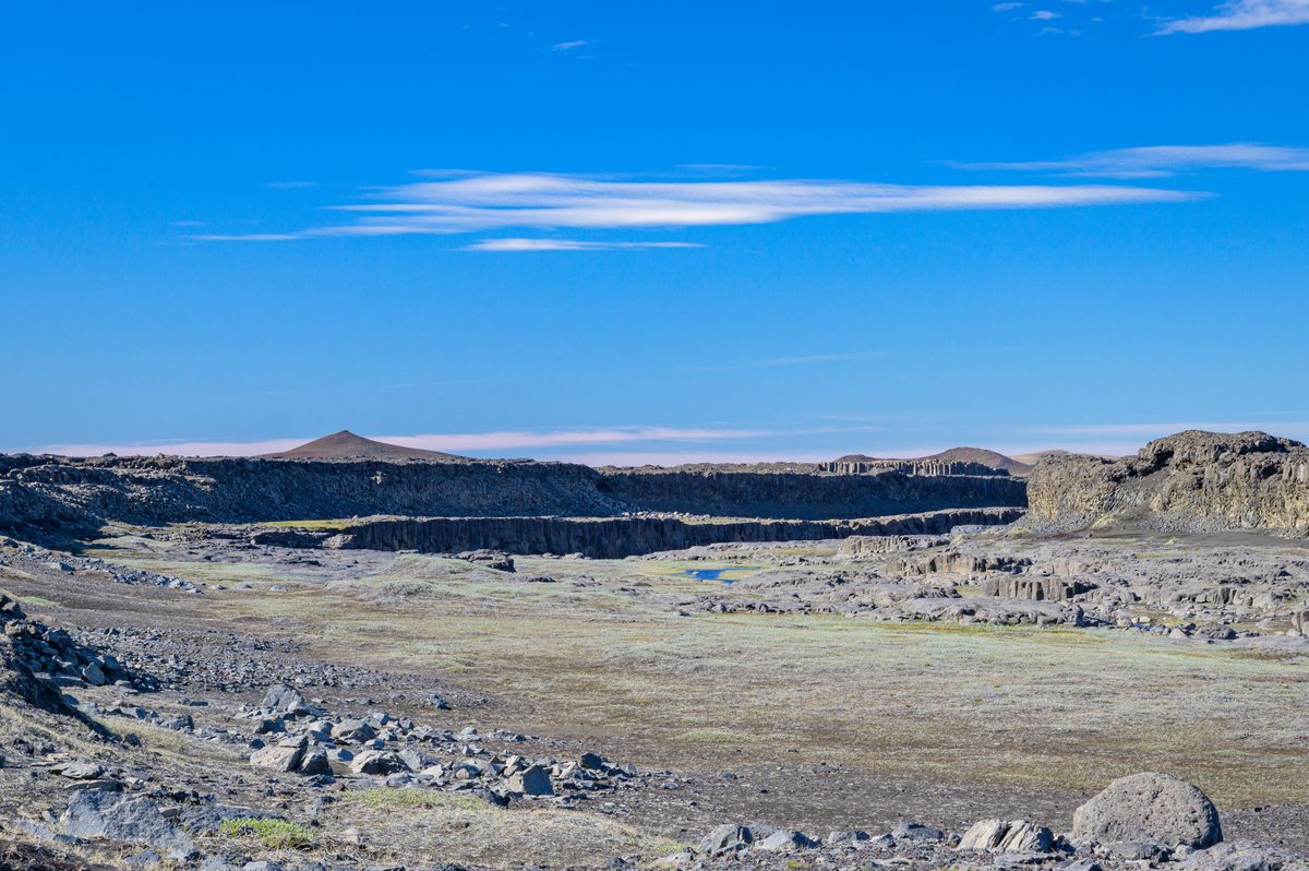 A long dark crater rim cuts across a pale, sparsely vegetated volcanic plain beneath a cloudless blue sky.