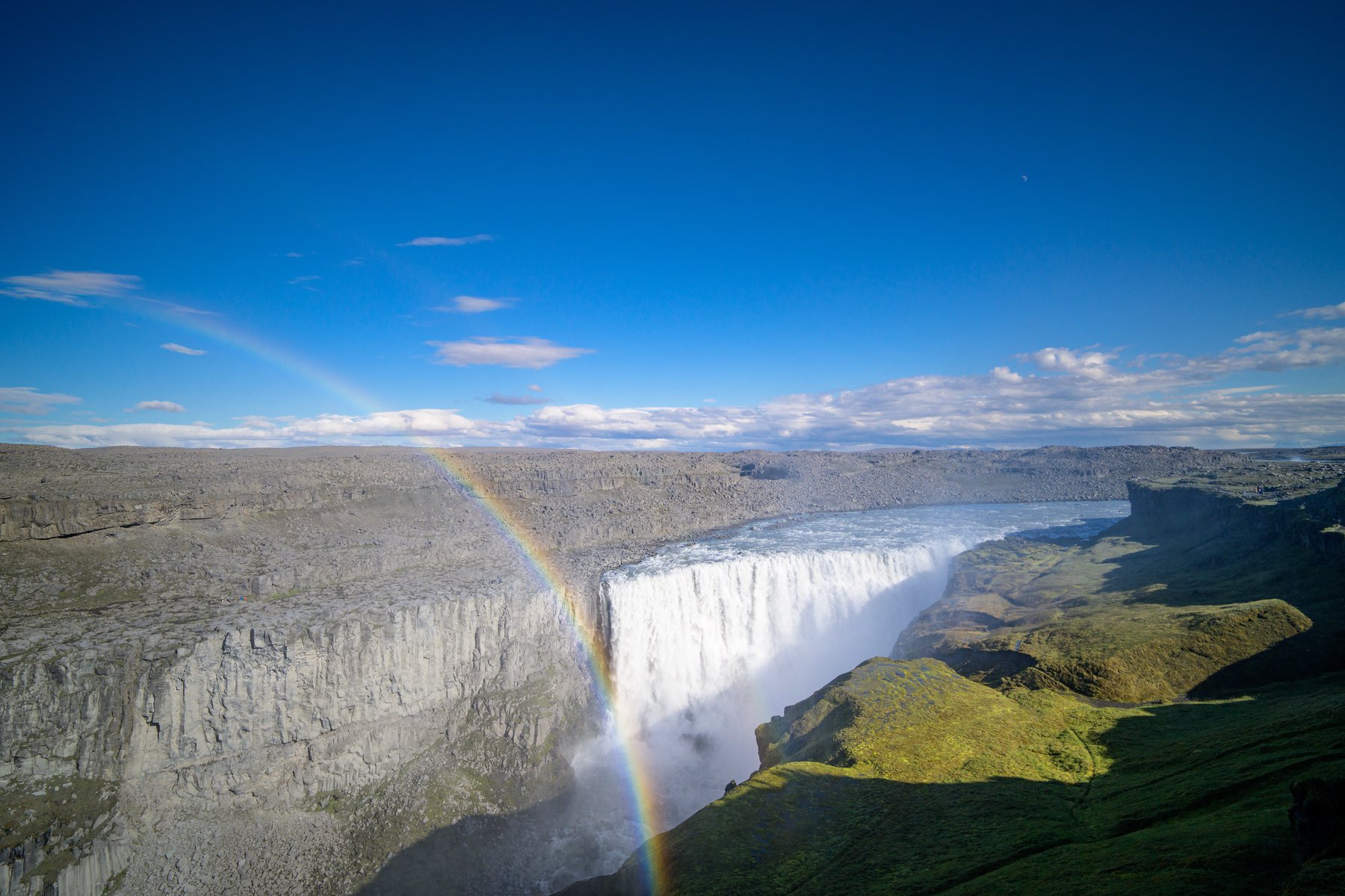 Dettifoss surges into its canyon while a rainbow arcs through the spray under a deep blue sky.