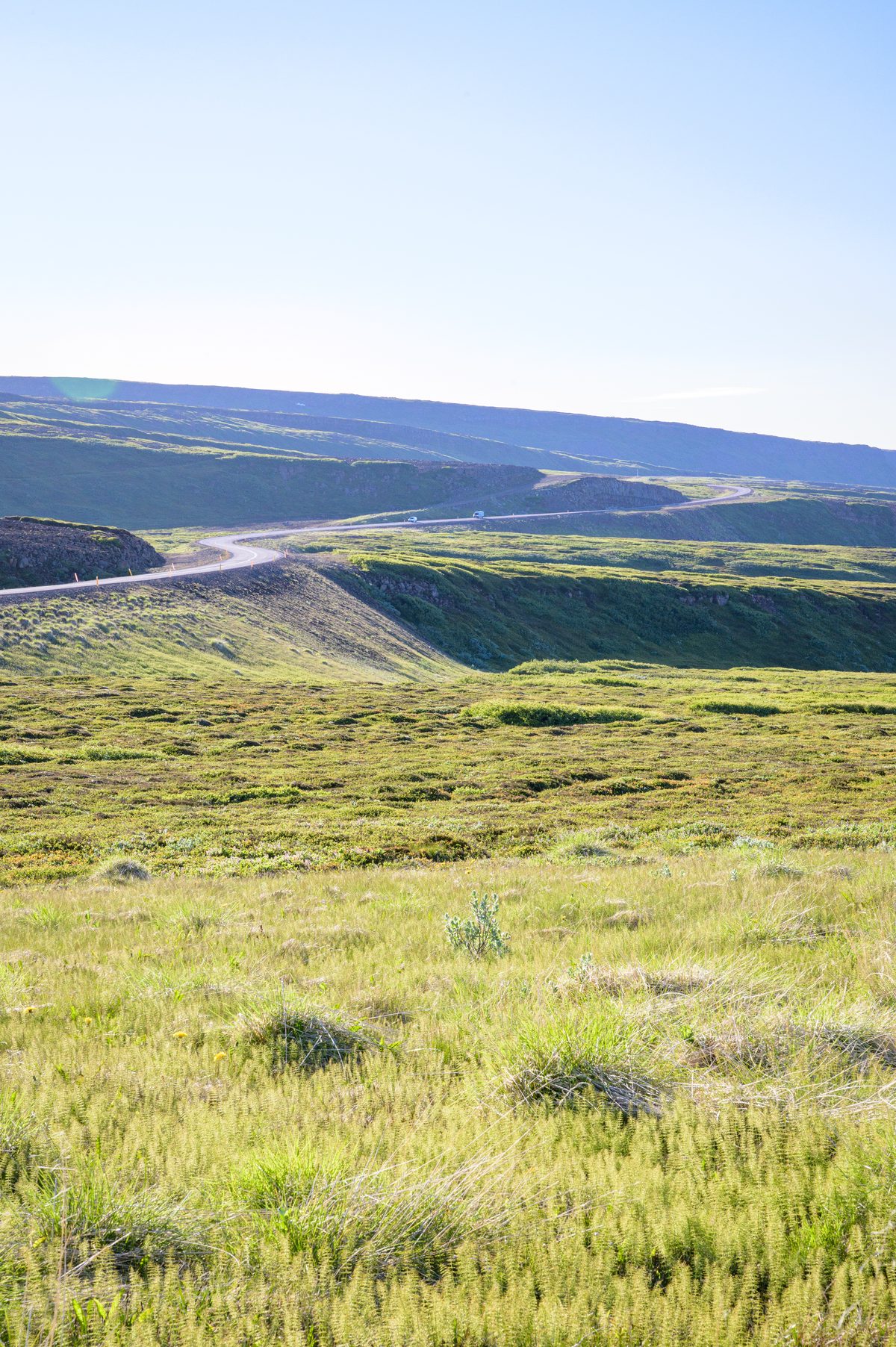 A narrow road winds across treeless green uplands toward layered hills under clear northern light.