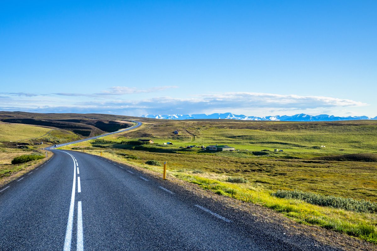 An empty two-lane road rolls across green coastal heath toward distant snow-covered mountains.
