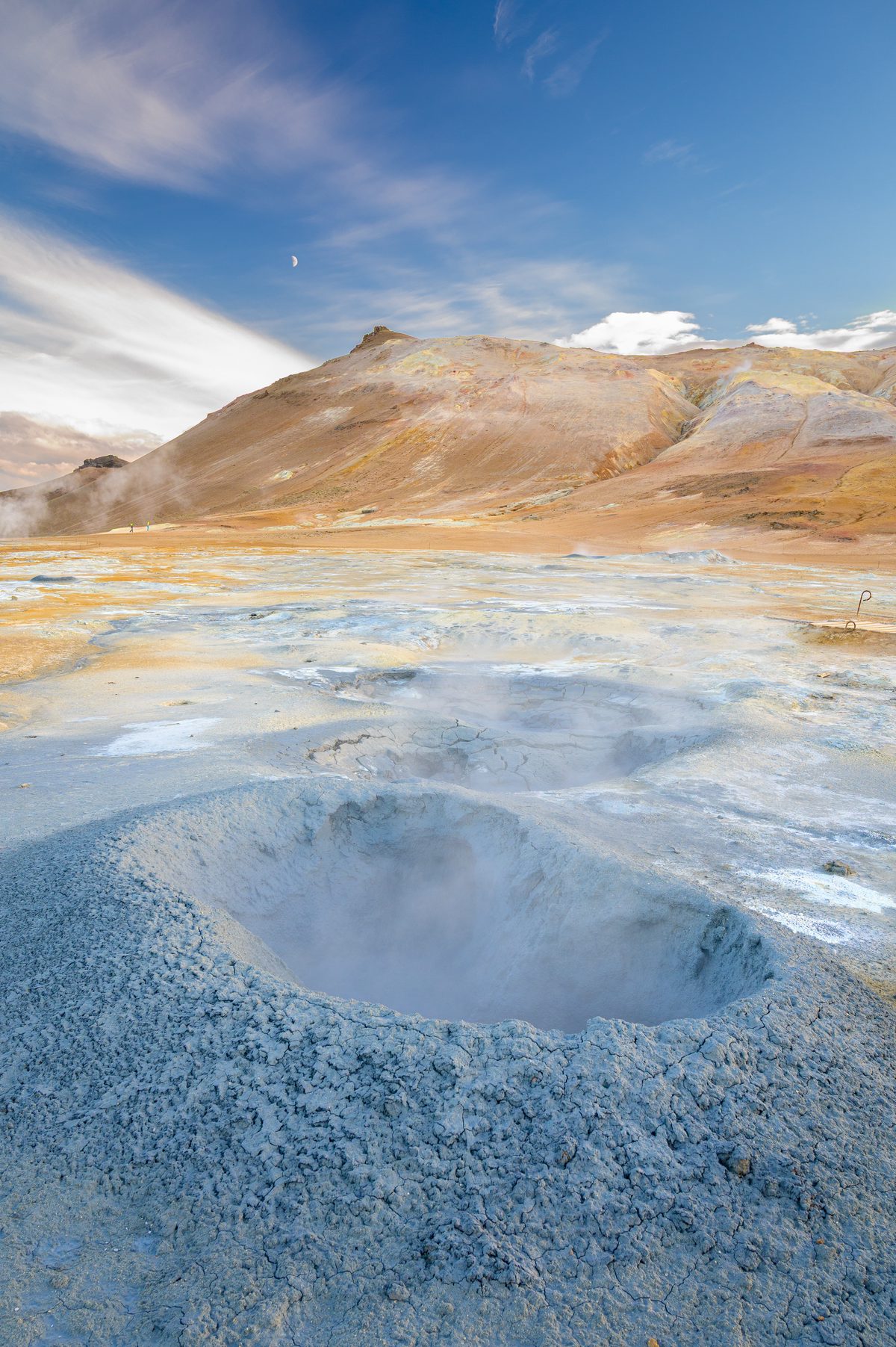 A vertical close view centers a pale blue mud pool in mineral-streaked ground below the ochre ridge.