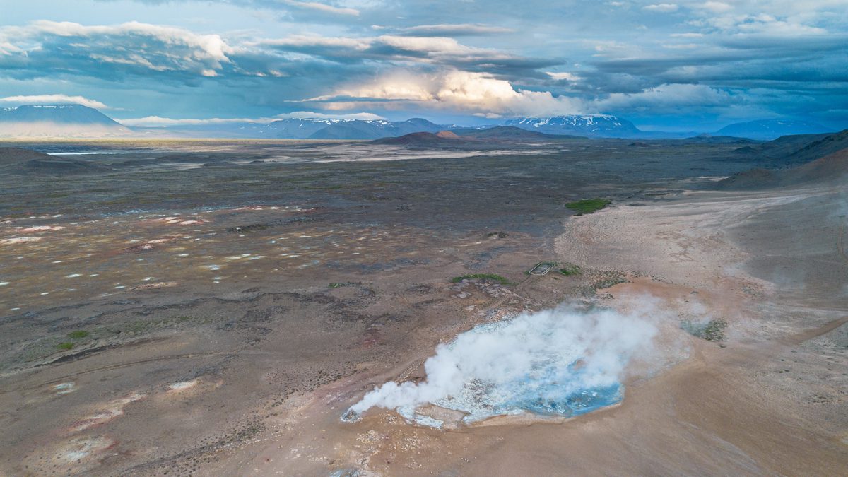 An aerial view looks across a pale blue geothermal vent releasing steam into the dark, sparsely vegetated volcanic plain of Krafla.