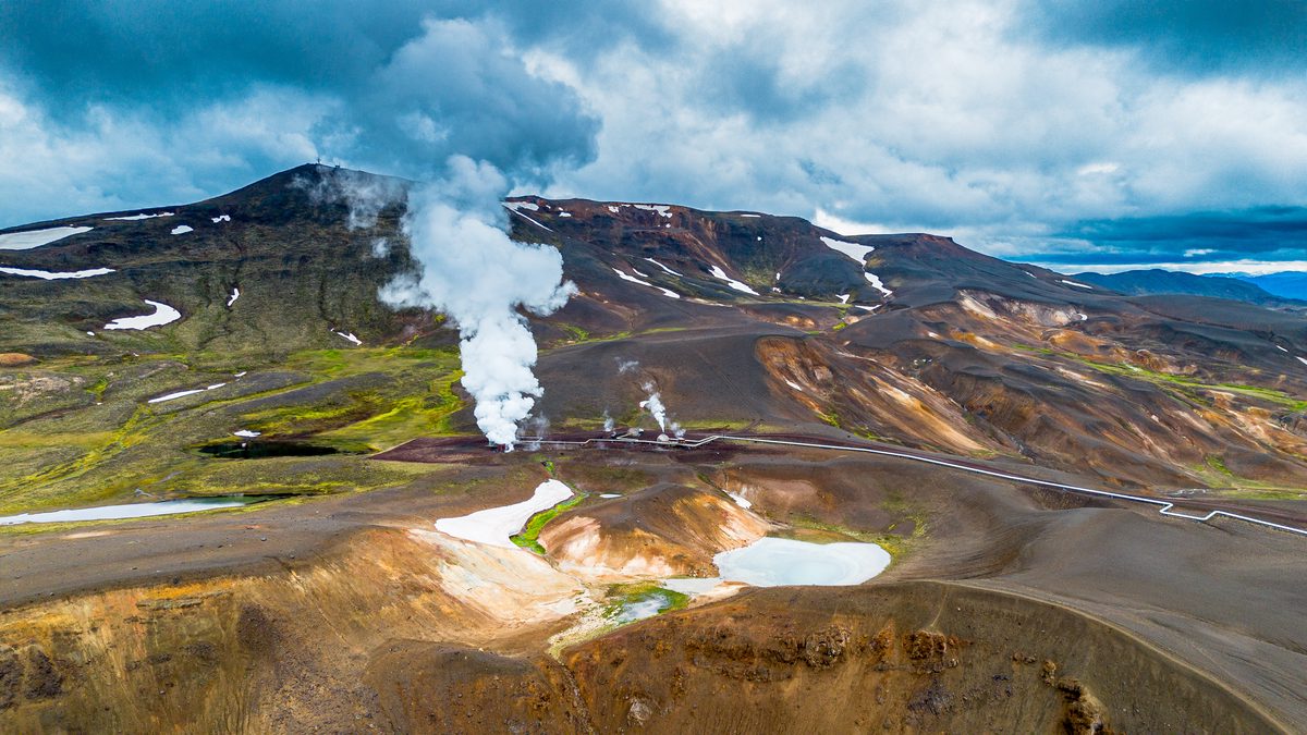 White steam rises from rust, ochre, and green geothermal slopes around winding roads in the Krafla volcanic area.