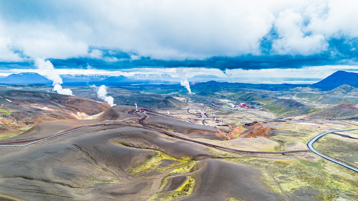 An aerial panorama reveals several steam plumes, mineral-colored ridges, service roads, and distant Lake Mývatn beneath low cloud.
