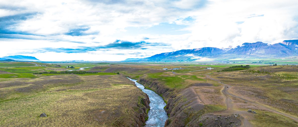 A narrow stream winds through a grassy hollow toward sunlit hills beneath the pale evening sky near Vatnsskarð.