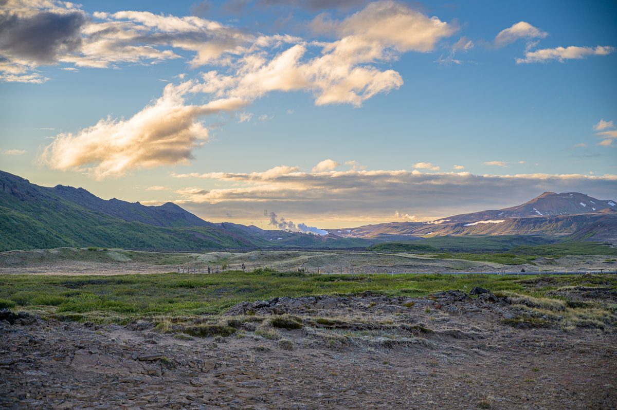 A wide volcanic valley stretches toward layered mountains beneath warm broken clouds and late northern light.
