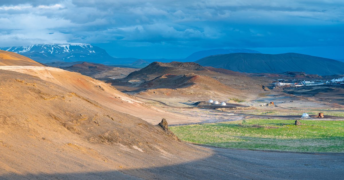 A panoramic view crosses orange geothermal hills toward dark lava, drifting steam, and distant snow-striped mountains.
