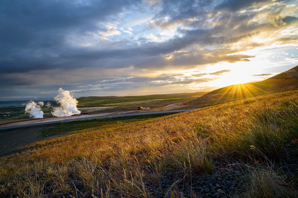 The low sun breaks beneath heavy clouds beside a geothermal steam plume, lighting tawny grass and dark volcanic slopes.