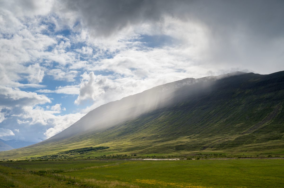 A veil of low white cloud pours across the dark crest and sunlit green face of a northern mountain.