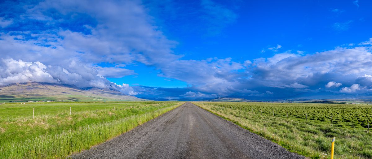 A straight gravel road divides a wide green plain and leads toward distant blue mountains beneath a sweeping sky.