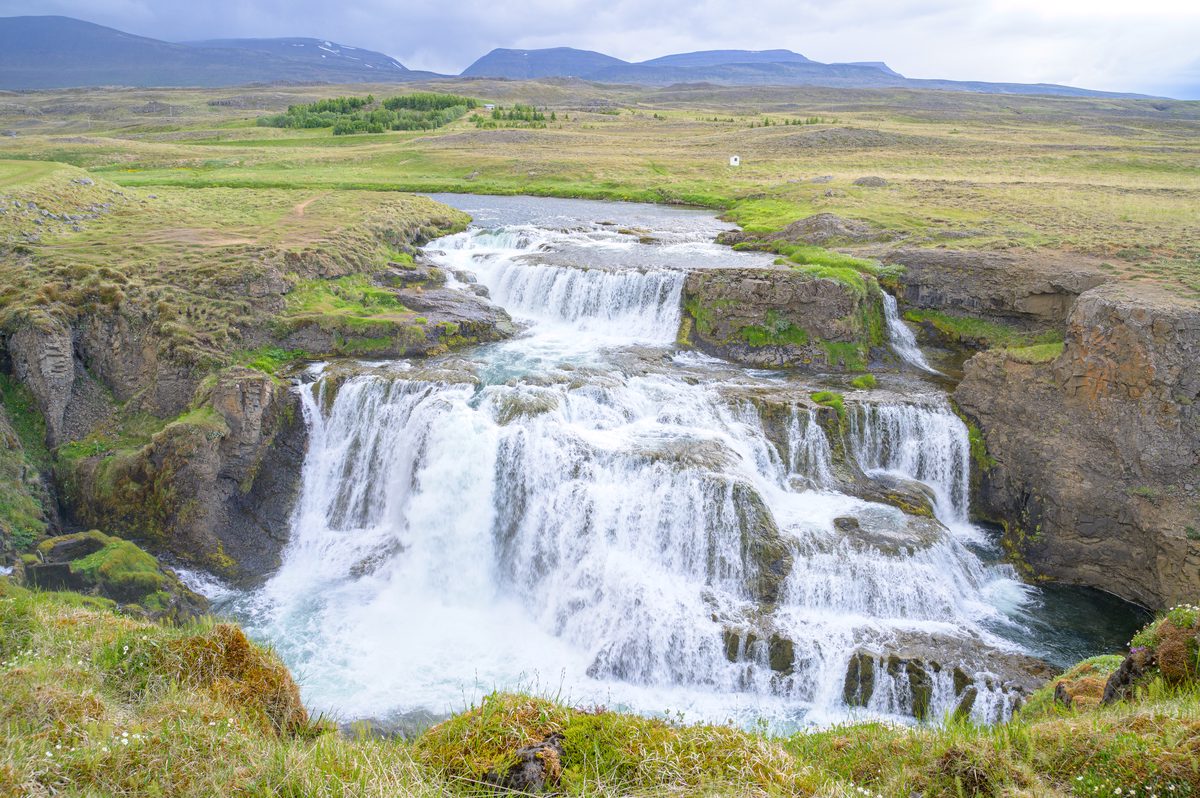 A clear river tumbles over several broad basalt ledges into a blue-green pool amid open grassland and distant mountains.