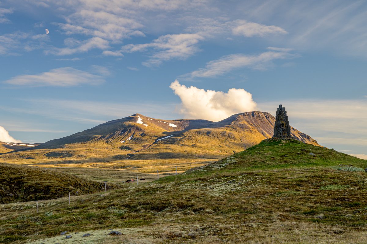 Minnisvarði um Stephan G. Stephansson stands on a grassy mound at Arnarstapi in Skagafjörður, before golden mountains, a bright cloud, and a pale moon.