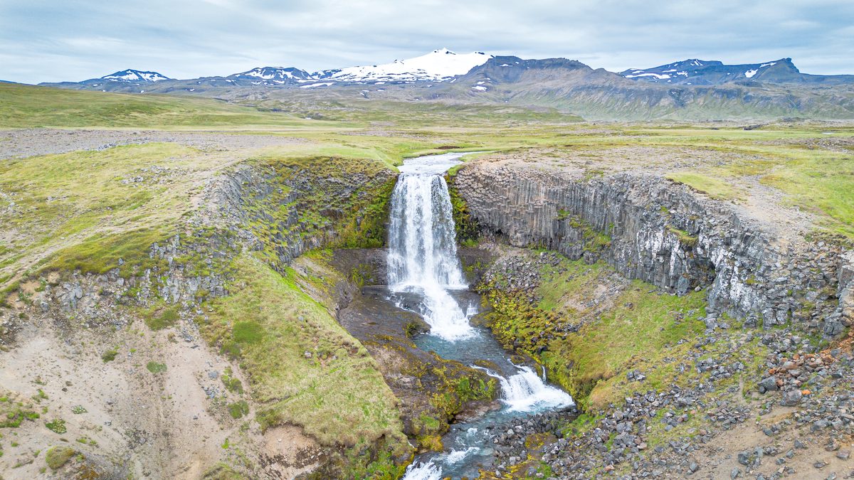 An aerial view of Svöðufoss shows white water dropping over a curved wall of basalt columns, with green plains and snow-covered Snæfellsjökull beyond.