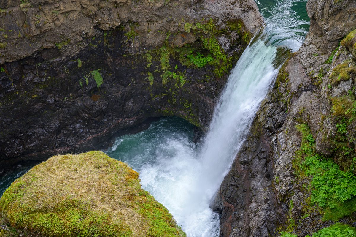 A close view into Kolugljúfur follows a narrow waterfall plunging between dark rock walls into a turquoise pool edged with moss and low plants.