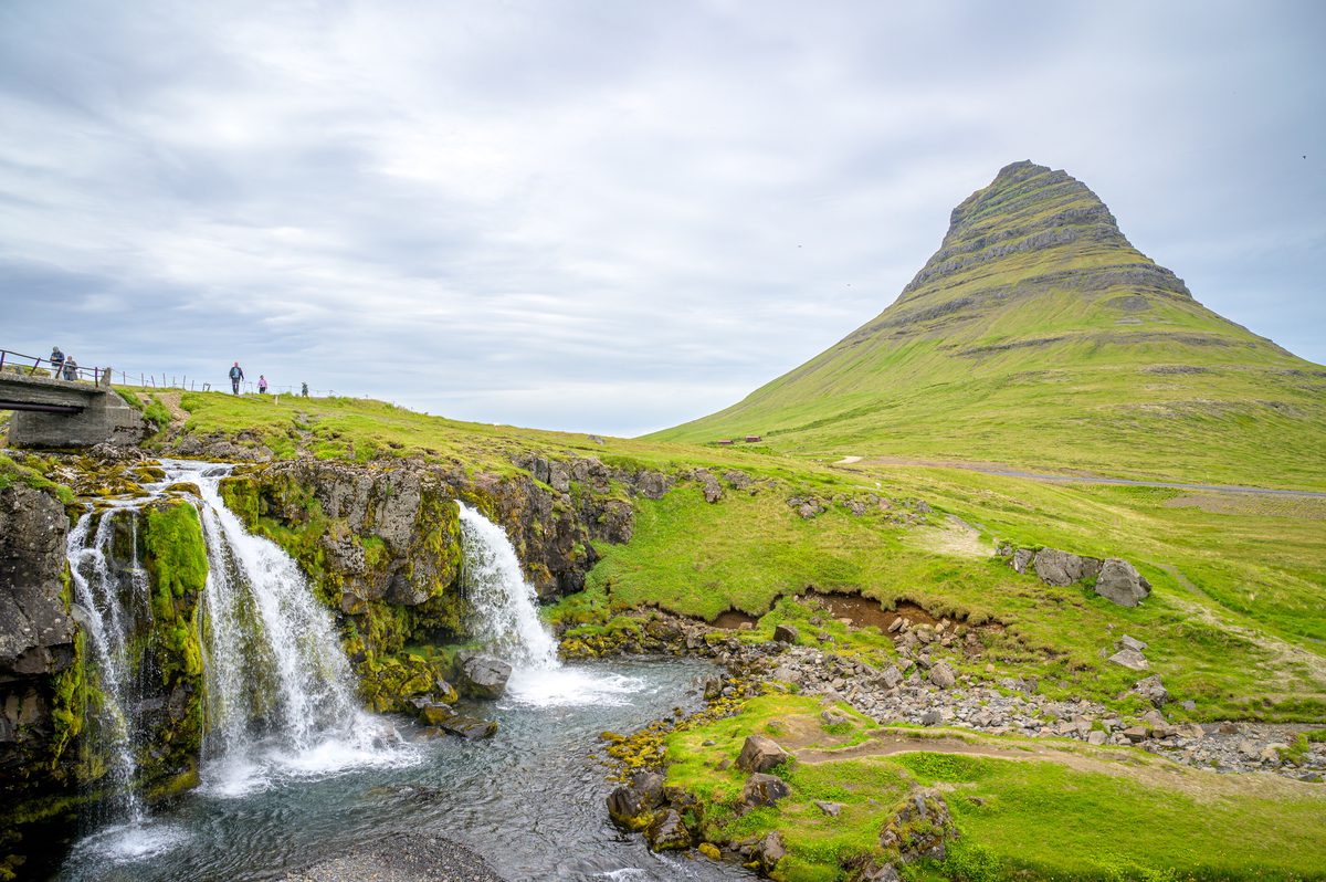 The cascades of Kirkjufellsfoss tumble over mossy rock in the foreground while the steep, layered peak of Kirkjufell rises across the green landscape.