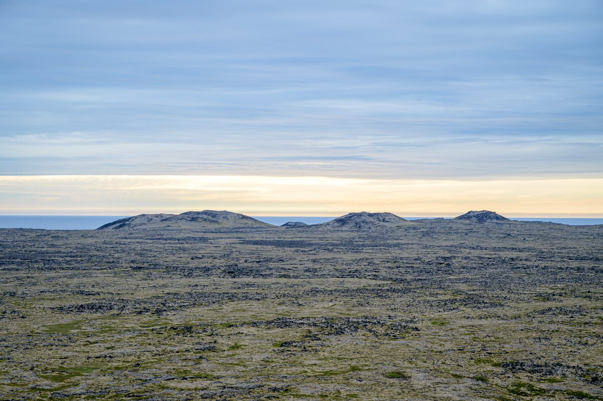 A row of low volcanic mounds breaks the horizon beyond an expansive, moss-covered lava field, under blue-gray clouds and a pale golden band of light.