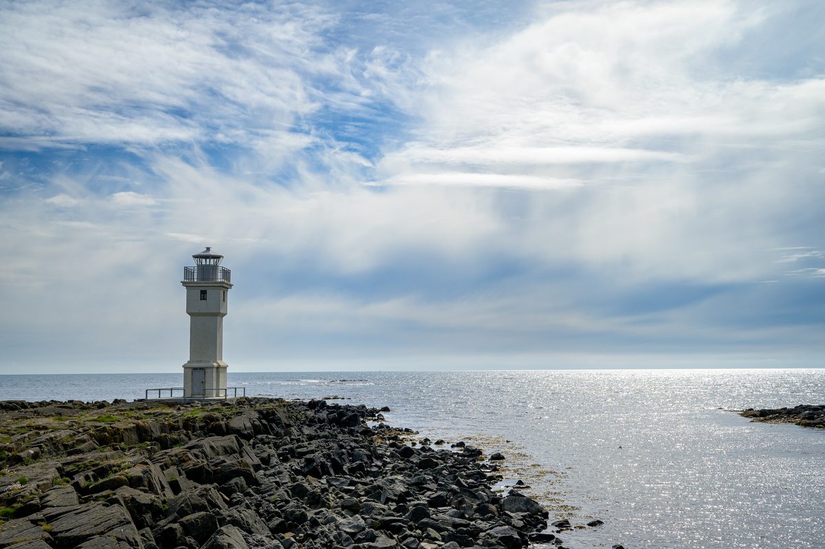 The old Akranes lighthouse stands on a dark rocky point beside sunlit water, beneath sweeping veils of white cloud.