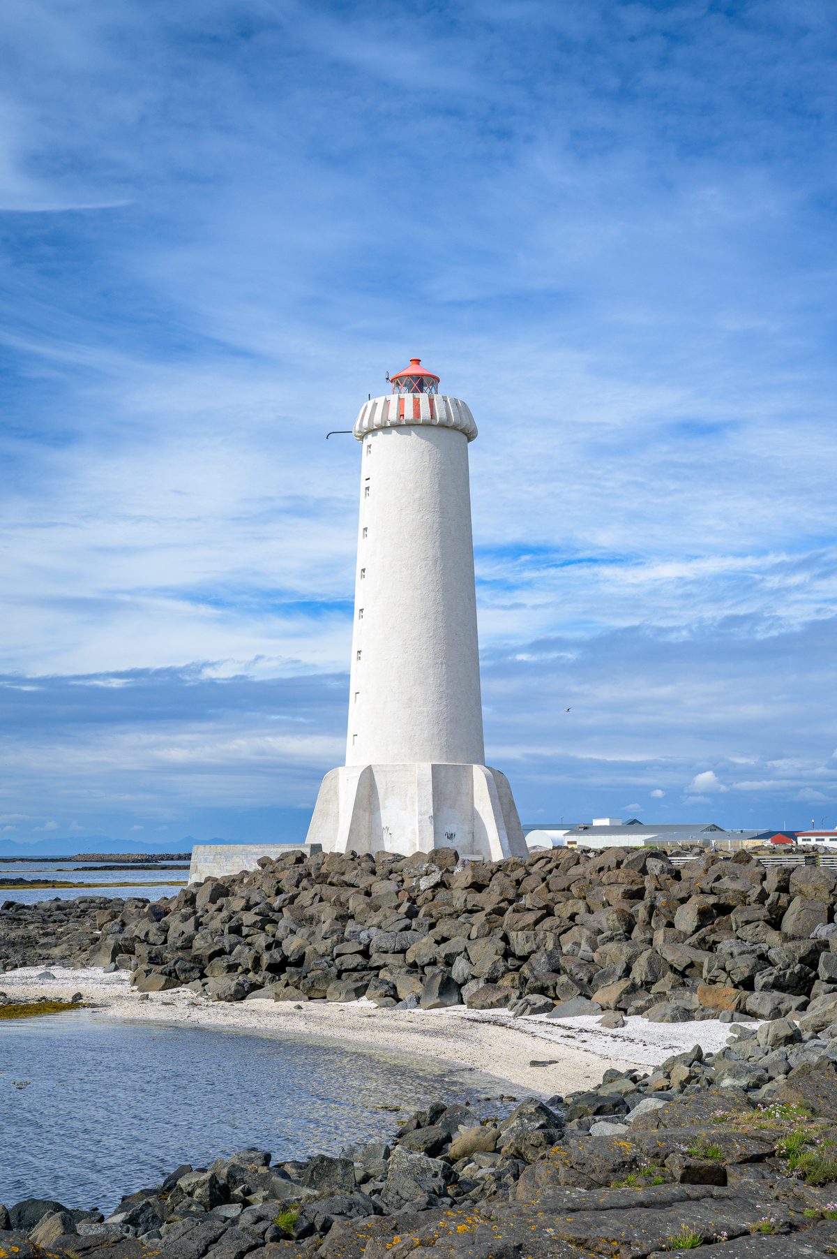 The tall white Akranes lighthouse rises above a rocky seawall and a small pale beach, framed vertically against a vivid blue sky.