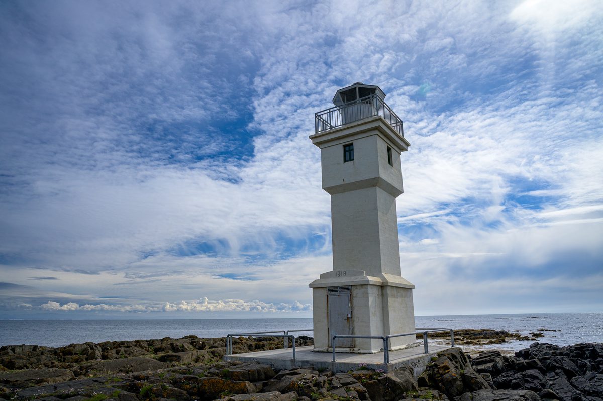 A close view of the old Akranes lighthouse shows its square white tower and glass lantern above dark coastal rocks beneath rippled clouds.