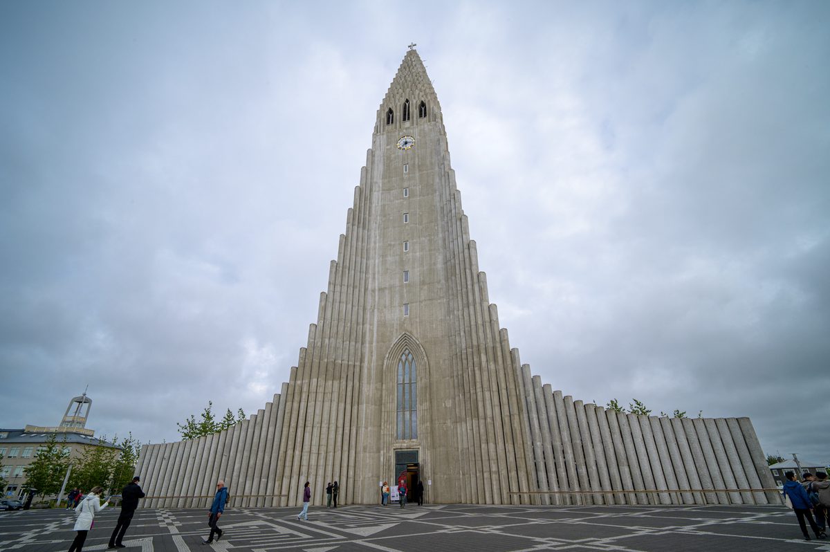 The stepped concrete façade and tall central spire of Hallgrímskirkja rise above visitors crossing the paved square in Reykjavík.
