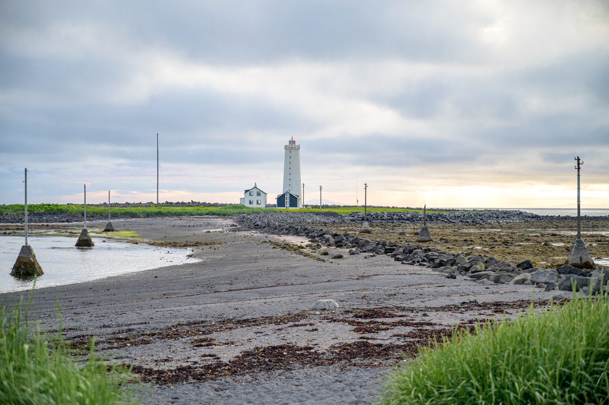 A white lighthouse and small buildings stand beyond a low-tide shore crossed by rocks and slender poles, while a band of warm light glows beneath gray clouds.