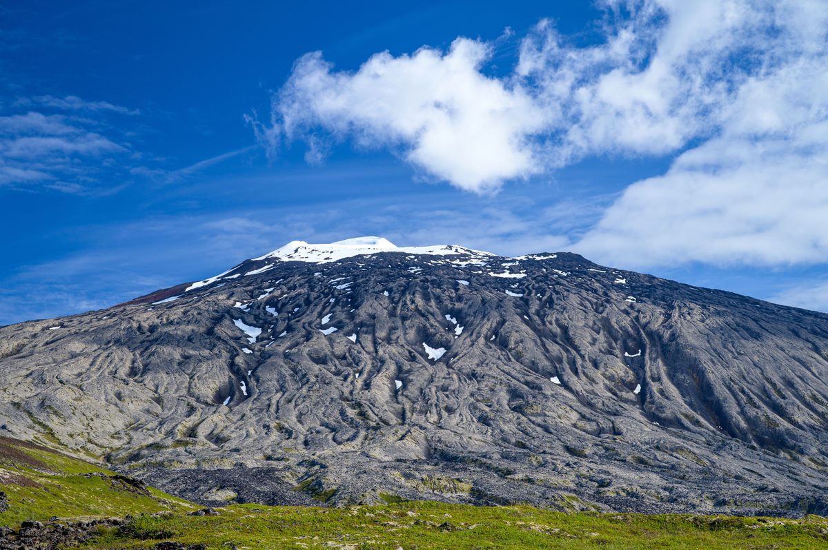 A broad mountain face is patterned with pale gullies and dark rock, rising from green ground to a snow-lined summit beneath a clear blue sky.