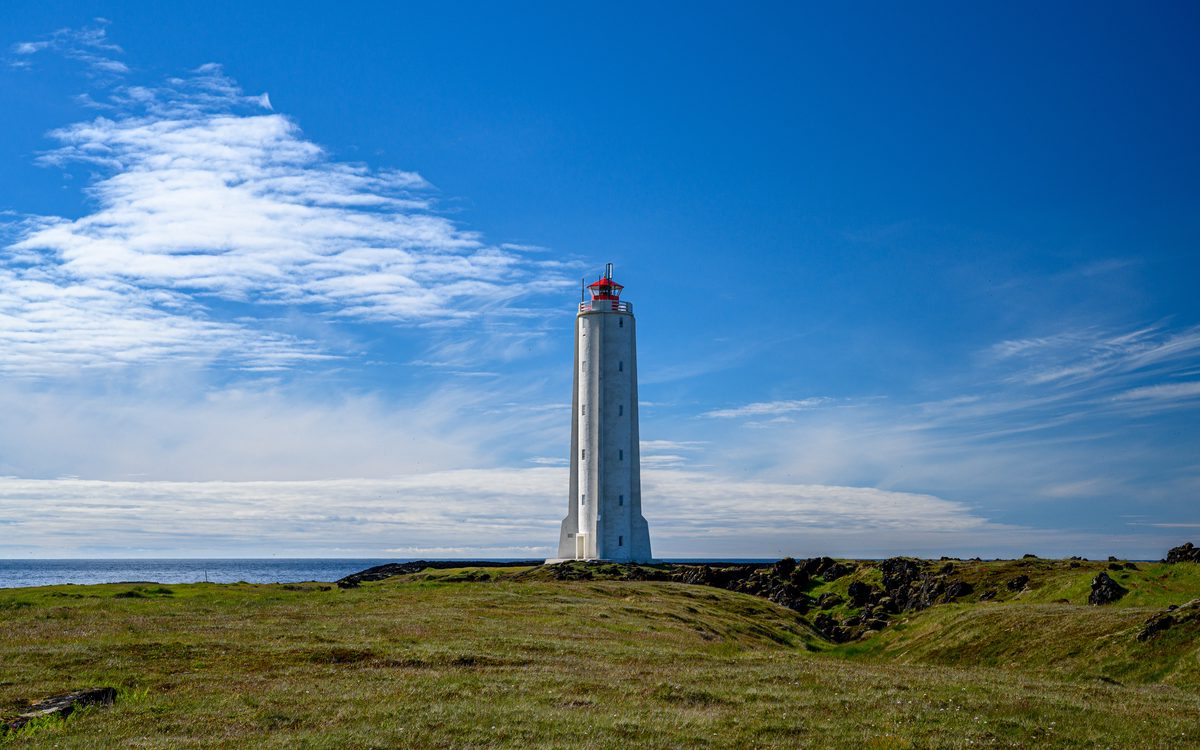 A slender white lighthouse with a red lantern stands on low green coastal ground beneath a deep blue sky brushed with white clouds.