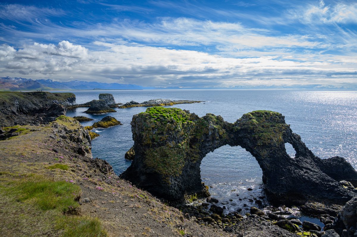 The dark rock arch of Gatklettur frames the water below, with green-topped cliffs and small sea stacks stretching along the coast at Arnarstapi.