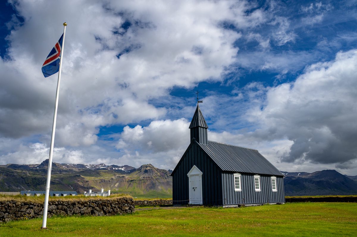 An Icelandic flag flies beside Búðakirkja, with the black church set on a green lawn against rugged mountains and sunlit clouds.