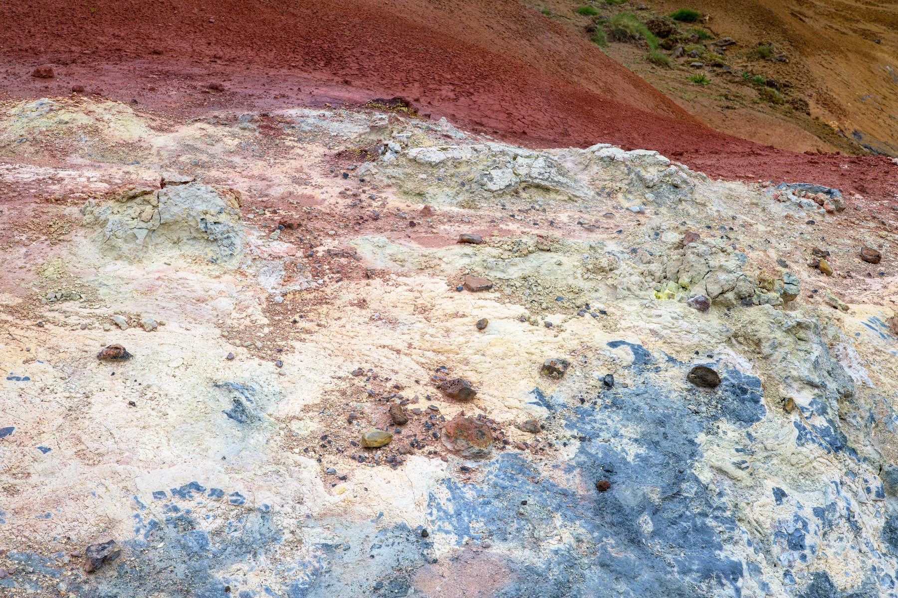 Blue-gray, cream, and yellow mineral crusts surround scattered stones at Seltún, contrasting with the deep red hillside above.