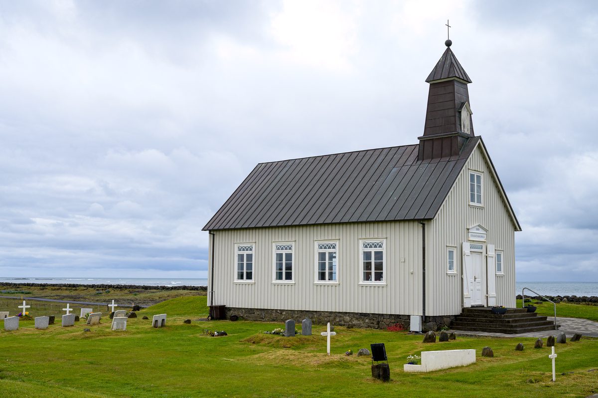 The pale timber walls and dark steeple of Strandarkirkja rise above a green churchyard dotted with gravestones and white crosses, with the Atlantic behind.