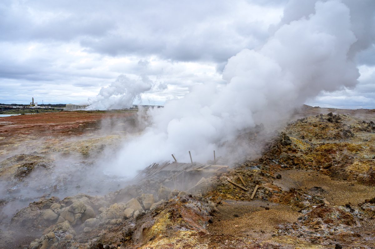 A dense plume of steam rises from Gunnuhver beside the broken remains of a wooden walkway, with mineral-stained rocks in the foreground and geothermal buildings on the horizon.